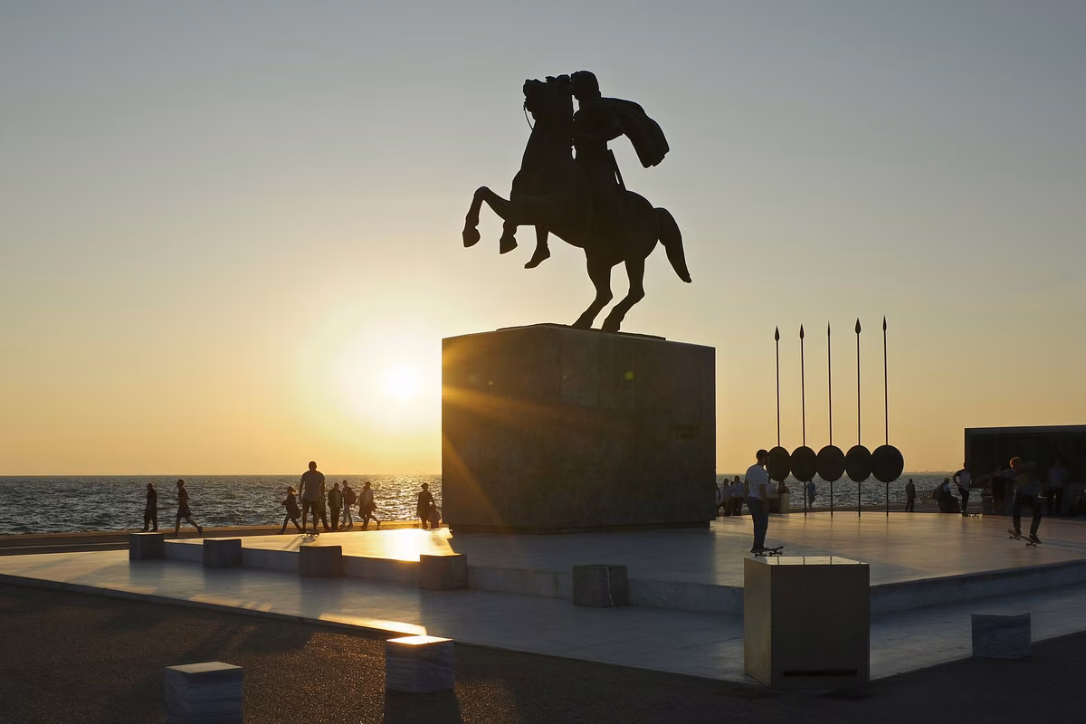 An equestrian statue in silhouette against the setting sun over the sea, with tall spears beside it