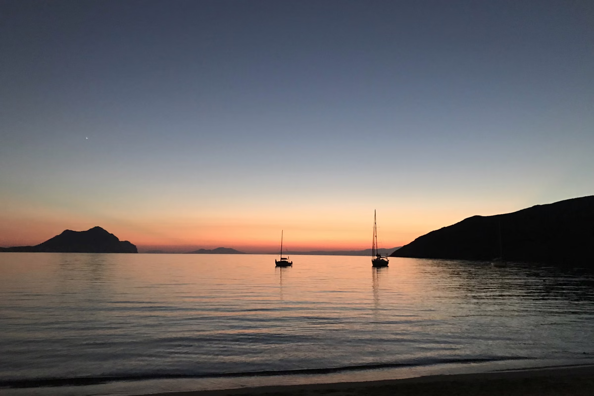 Sunset over a calm bay with two anchored sailing boats and the dark outline of an islet