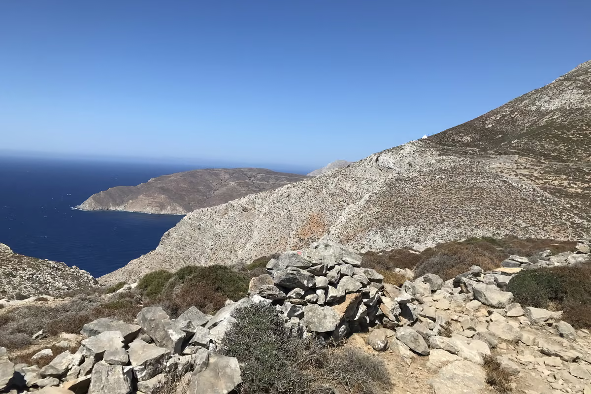 A rocky footpath on a bare hillside high above a deep blue sea, with a headland and a white chapel on the ridge