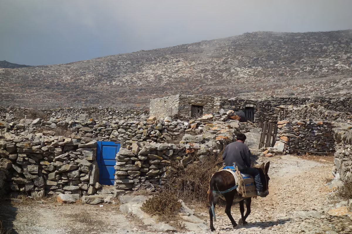 A man riding a donkey past low stone houses with a blue door, under a bare, rocky hillside