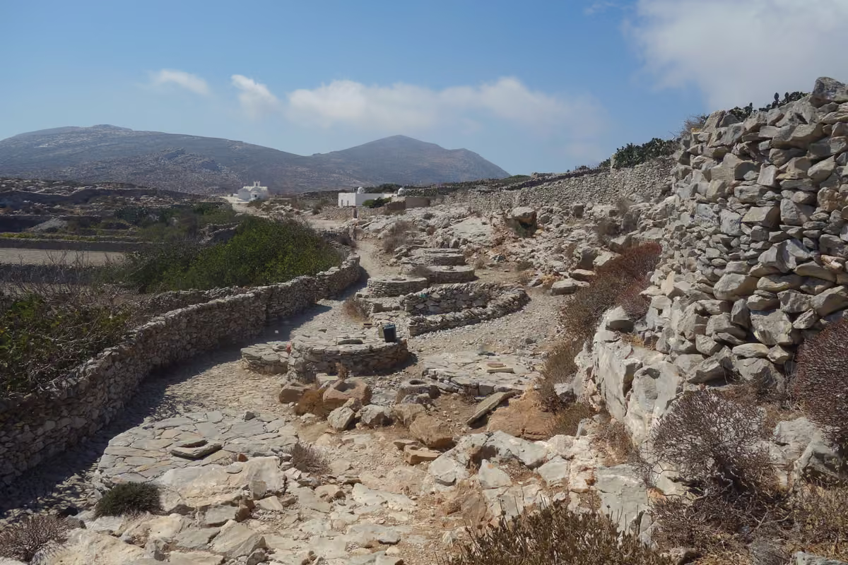 An old stone-paved path between dry-stone walls, climbing through rocky ground towards white chapels and bare hills