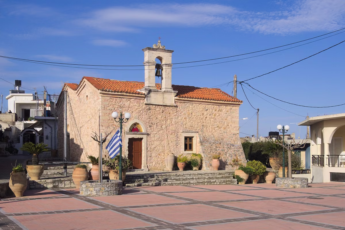 A stone church with a bell gable on a paved village square, big clay jars around it
