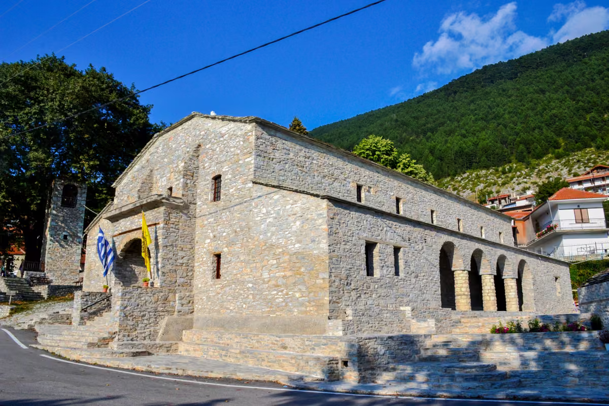 A stone church with a bell tower and arcade on a village street, a wooded slope behind