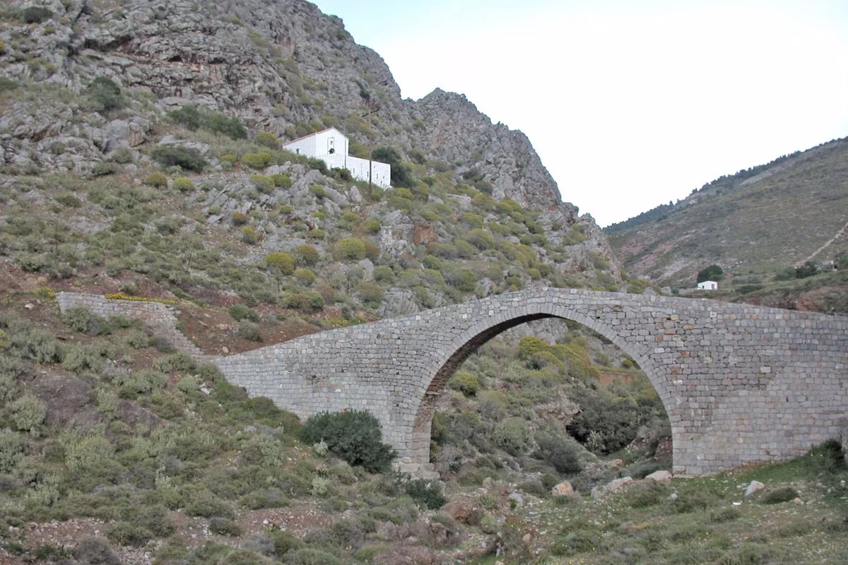 A single-arch stone bridge on a bare hillside above Palamidas, with a white chapel behind it