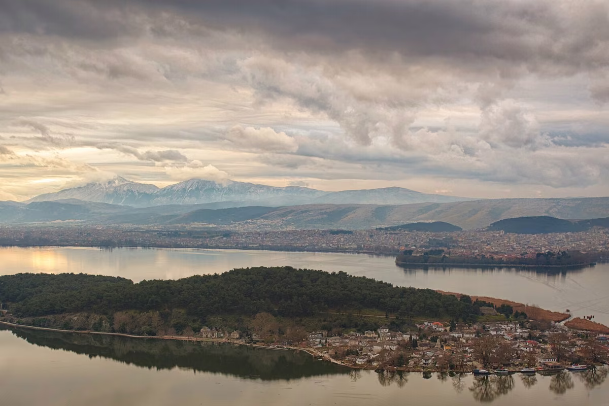 A wooded island in a calm lake at dusk, snowy mountains behind