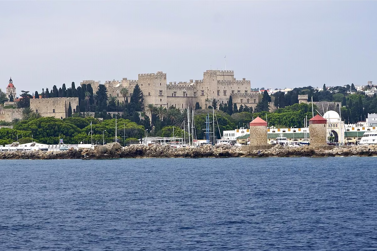 A walled medieval palace above a harbour with windmills, seen from the sea