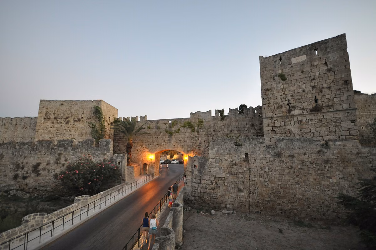 A lit gateway in massive medieval walls reached by a bridge, at dusk
