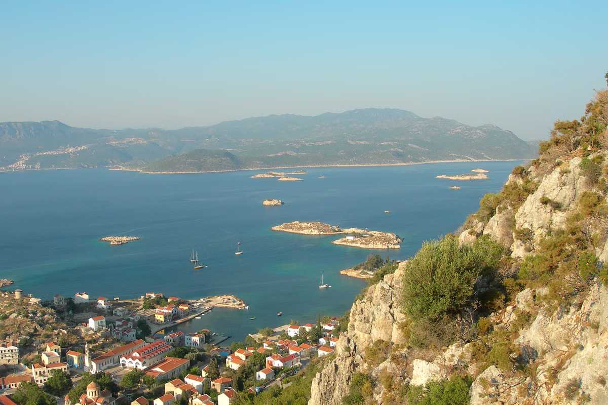 Red roofs of a harbour town far below a rocky slope, with islets in a turquoise sea and a mountainous coast beyond